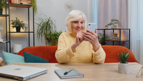 Eine-ältere-Großmutter-Nutzt-Zu-Hause-Ihr-Smartphone-Und-Feiert-Den-Erfolg,-Den-Sieg-Und-Das-Siegreiche-Online-Spiel