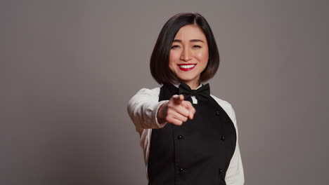Asiatische-Kellnerin-Zeigt-Auf-Die-Kamera,-Um-Sie-Vor-Grauem-Hintergrund-Auszuwählen