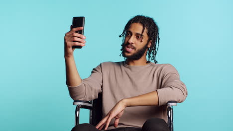 Man-in-wheelchair-saluting-friend-over-online-videocall-on-cellphone,-studio