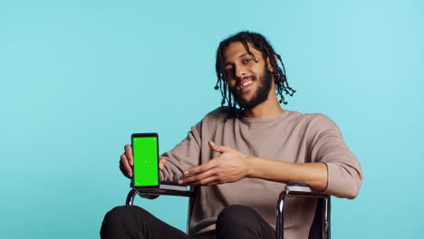 Man-in-wheelchair-pointing-towards-green-screen-mobile-phone,-studio-background