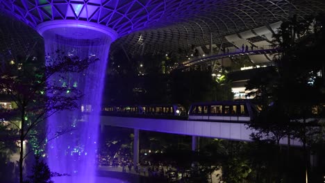 Lila-Beleuchteter-Indoor-Wasserfall-Am-Jewel-Changi-Airport-Singapur-Am-Abend-Mit-Vorbeifahrendem-Skytrain