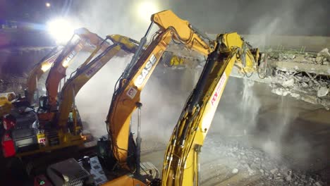 An-aerial-truck-pan-of-a-team-of-contractors-demolishing-a-reinforced-concrete-bridge-with-jackhammer-excavators-at-night,-Canada