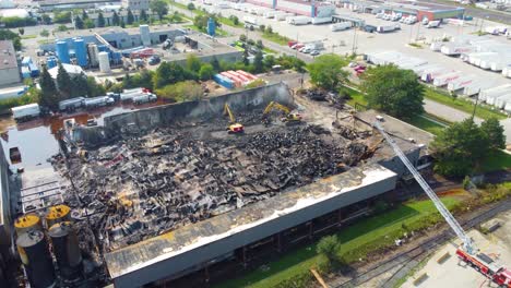 Aerial-drone-shot-descending-on-the-remains-of-a-propane-depot-after-an-industrial-fire,-as-a-team-of-firefighters-with-excavators-clear-the-debris-at-the-devastating-scene,-Canada