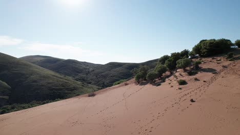 Aerial-dolly-above-sloping-sand-dune-with-grassy-mountain-side-behind,-footsteps-across-landscape