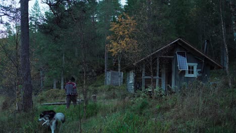 Man-With-His-Dog-Walking-To-The-Old-Wooden-Cabin-On-A-Windy-Morning