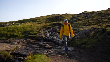 Male-hiker-in-yellow-jacket-crosses-stream-in-Leitisvatn,-Faroe-Islands