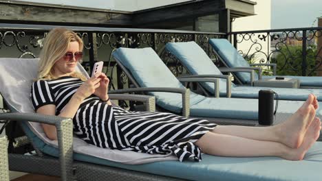 Blonde-fashion-girl-vibing-to-music-lying-on-a-rooftop-deck-chair-at-day-time-with-Bluetooth-speaker-next-to-her