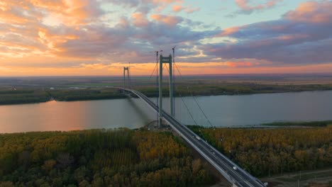 Aerial-close-up-shot-showcasing-a-suspended-bridge-spanning-a-vast-river,-cars-driving,-after-sunset-orange-colored-sky,-4k50fps
