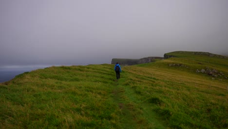 Male-hiker-walk-along-grassy-hill-on-Mykines-and-turns-back-calling-for-someone-on-a-cloudy-day,-Faroe-Islands