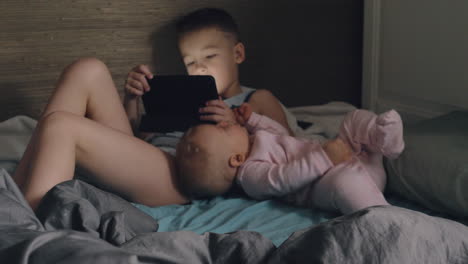 Ein-Junge-Mit-Einem-Tablet-Und-Seiner-Kleinen-Schwester-Auf-Einem-Unordentlichen-Bett