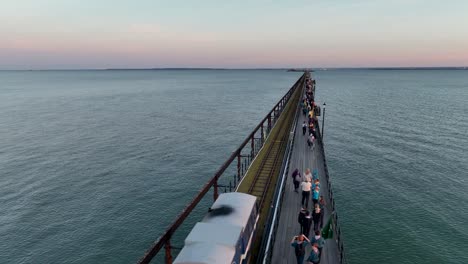 Southend-Pier-Sunset-drone-pivot-shot-shows-people-walking-along-pier-as-pier-train-enters-shot-and-travels-along-rails,-then-pulls-back-and-follows-train