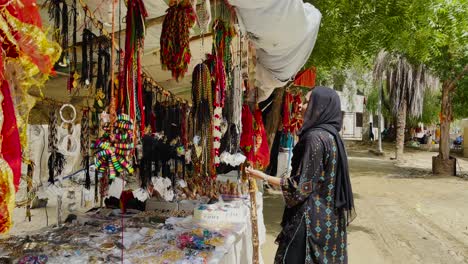 Pakistani-Woman-Wearing-Head-Scarf-Walking-Towards-Market-Stall-Selling-Colourful-Beads-In-Hingol,-Balochistan