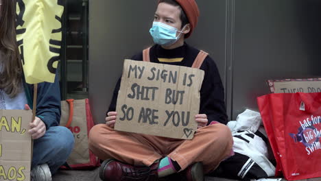 A-masked-protestor-wearing-an-orange-beanie-and-dungarees-sits-crosslegged-holding-a-cardboard-placard-that-reads,-“My-sign-is-shit-but-so-are-you”