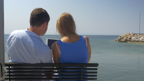 Young-woman-and-man-are-sitting-on-bench-on-beach-on-sea-skyline-background-watching-something-in-tablet-computer-and-speaking-Piraeus-Greece