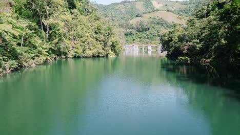 Aerial-approaching-shot-of-green-river-with-dam-of-tireo-during-sunny-day,-Dominican-Republic