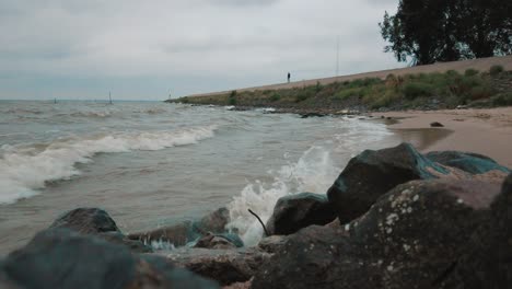 Overlooking-waves-crash-into-beach,-man-stands-in-the-background-watching-the-lake