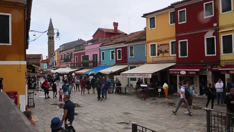 Tourist-stroll-Burano-street-of-colourful-houses,-look-at-souvenir-shops-on-rainy-day,-Venice,-Italy