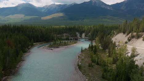 Descending-drone-shot-of-the-turquoise-glacial-waters-of-the-Kootenay-river-on-a-sunny-summer-day-with-a-bridge-in-view-and-logging-roads-with-mountains-in-the-background