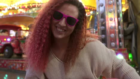 woman-with-smile-and-sunglasses-dances-looking-at-camera-with-fairground-background-behind-her