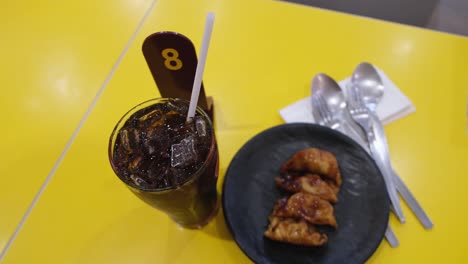Cola-drink-in-a-glass-with-ice-and-some-dumplings-on-a-small-dish-with-a-queue-number-eight,-two-pair-of-spoon-and-fork