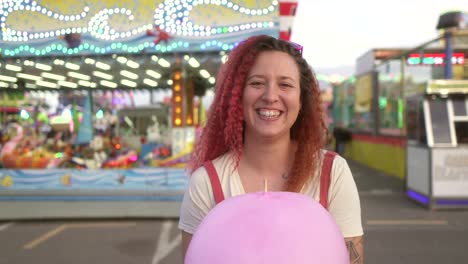 happy-woman-with-red-hair-smiles-at-camera-with-cotton-candy-and-fair-background