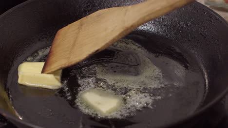 Wooden-spatula-stirs-melting-butter-in-hot-cast-iron-pan-to-coat-base