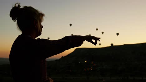 Eine-Silhouettierte-Frau-Zeigt-Mit-Heißluftballons-Bei-Sonnenaufgang-Am-Frühen-Morgen-In-Den-Himmel