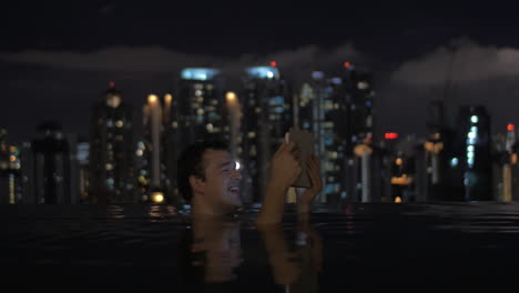 Blick-Auf-Einen-Mann-Im-Schwimmbad-Auf-Dem-Dach-Eines-Wolkenkratzers-Mit-Tablet-Vor-Der-Nächtlichen-Stadtlandschaft-Von-Kuala-Lumpur,-Malaysia