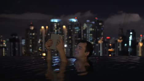 Im-Pool-Auf-Dem-Dach-Eines-Hotels-In-Kuala-Lumpur,-Malaysia,-Schaut-Sich-Ein-Mann-Ein-Video-Auf-Einem-Tablet-An-Und-Lächelt