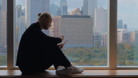 Mujer-Joven-Sentada-En-El-Alféizar-De-La-Ventana-Usando-Una-Tableta-Y-Mirando-La-Ventana-Hong-Kong,-China