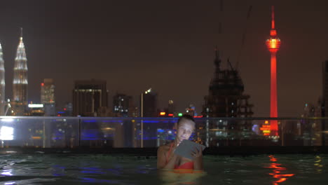 Blick-Auf-Eine-Frau-Im-Schwimmbad-Auf-Dem-Dach-Des-Wolkenkratzers-Mit-Tablet-Vor-Der-Nächtlichen-Stadtlandschaft-Von-Kuala-Lumpur,-Malaysia