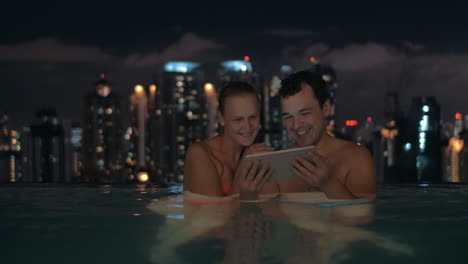 Nachtansicht-Eines-Jungen-Schönen-Paares-Im-Schwimmbad,-Das-In-Einem-Tablet-Auf-Dem-Dach-Eines-Wolkenkratzers-Und-Der-Stadtlandschaft-Kuala-Lumpur-Malaysia-Zuschaut