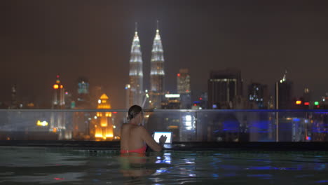 View-of-woman-in-swimming-pool-on-the-skyscraper-roof-using-tablet-against-night-city-landscape-Kuala-Lumpur-Malaysia