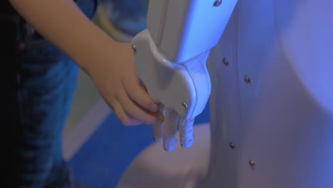 Vista-De-Cerca-De-La-Mano-Del-Niño-Sostiene-La-Mano-De-Plástico-Blanco-De-Un-Robot-Humanoide-En-Una-Exposición-De-Robótica-En-Moscú,-Rusia