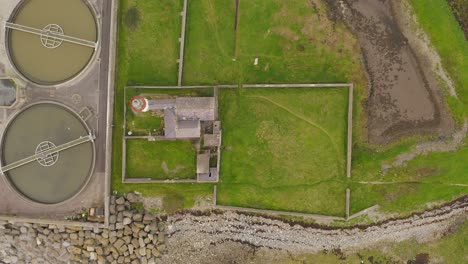 Old-lighthouse-next-to-waste-water-treatment-plant-on-grass-island,-aerial-top-down