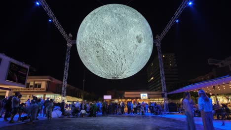 Large-bright-moon-art-exhibit-at-city-square-at-night,-people-taking-selfies-with-supermoon,-timelapse