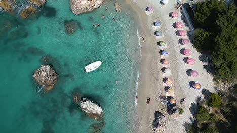 Top-down-drone-rises-as-ocean-waves-crash-gently-on-shoreline-with-umbrellas-and-boat-on-shore