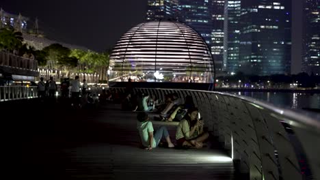 Menschen-Saßen-Neben-Dem-Geländer-Und-Chillten-Auf-Der-Promenade-Neben-Dem-Marina-Bay-Sands-Hotel-Mit-Blick-Auf-Den-Apple-Store-Im-Hintergrund-Bei-Nacht-In-Singapur