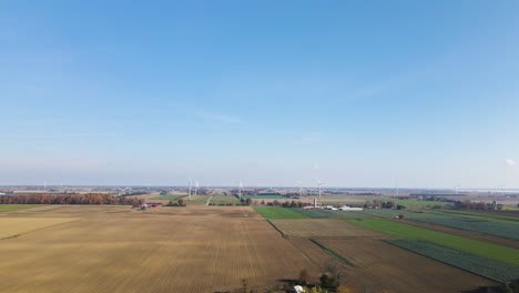 Aerial-pullback-above-farmland-crop-fields-with-wind-turbines-in-distance,-beautiful-blue-sky-above