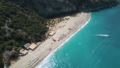 High-angle-bird's-eye-view-above-white-sandy-beach-in-the-Albanian-Riviera,-boat-drives-out-to-sea