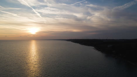 Panoramic-aerial-overview-above-dark-silhouette-of-land-as-sun-sets-onto-horizon-above-lake