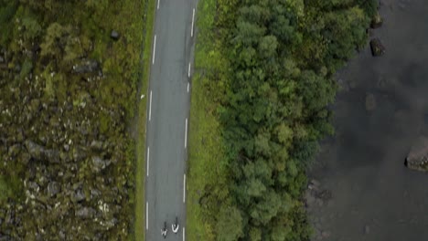 Aerial-drone-shot-of-cyclists-cycling-down-mountain-road-by-lake