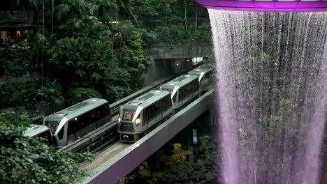 A-slow-motion-shot-of-the-cascading-water-of-the-Vortex-indoor-waterfall-at-the-Jewel-Changi-Airport-as-the-SkyTrain’s-pass-by-below,-Singapore
