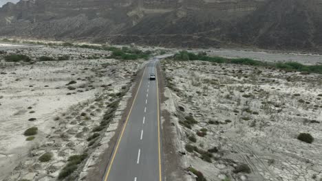 Aerial-drone-backward-moving-shot-over-a-car-moving-along-a-highway-in-Hingol-National-Park-with-mountain-range-in-the-background-in-Balochistan-on-a-cloudy-day