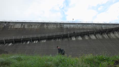 Low-Angle-View-Of-Male-Hiker-Waking-Past-Concrete-Dam-Wall