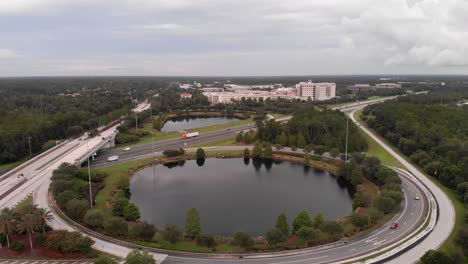Aerial-drone-shot-revealing-the-highway-and-roads-cars-passing-by-blue-sky-pond-white-clouds-trees-in-on-side-of-highway