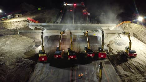 Aerial-view-over-machinery-demolishing-an-old-road-bridge,-at-night