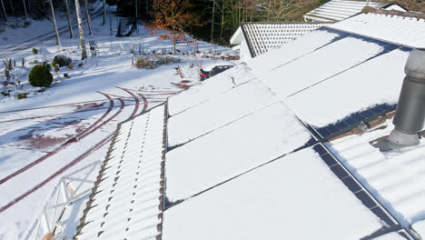 Aerial-view-backwards-low-over-solar-cells-on-a-snowy-house-roof,-winter-day