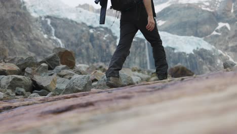 Tracking-Shot-Of-Male-Hiking-Over-Rocks-With-Reveal-Of-Snow-Covered-Mountains-In-Background