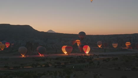 Beleuchtete-Heißluftballons-Starten-Am-Frühen-Morgen-In-Einer-Dunklen-Landschaft
